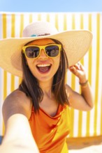 Young woman wearing sunglasses and a straw hat taking a selfie while smiling at the camera on a