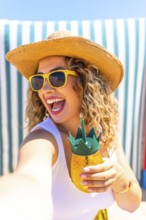 Happy tourist woman wearing sunglasses and straw hat taking a selfie at beach while holding a