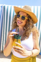 Young curly blonde woman wearing sunglasses and straw hat drinking juice at beach bar, enjoying