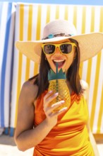 Young woman wearing sunglasses and a straw hat enjoying a refreshing pineapple juice at the beach