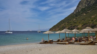Kamari beach, summer beach scenery with deckchairs and parasols, two sailing boats in the calm blue