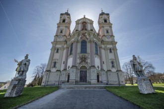 East tower of the baroque basilica backlit, in front the two church patrons, St Alexander on the
