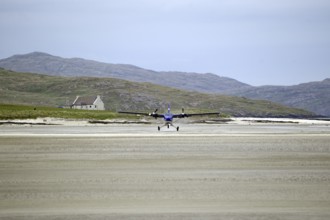 Aeroplane landing on a sandy runway on a beach with mountains and a building in the background,