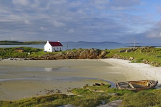 Boat wreck on a sandy beach with a white house and rocks in the background, Isle of Barra,