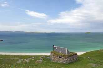 Stone cottage with grassy roof overlooking the vast sea and green hills under a blue sky, Eriksay,