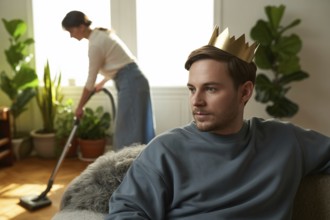 Man with golden crown relaxing on couch while woman vacuums in background. Concept of patriarchal