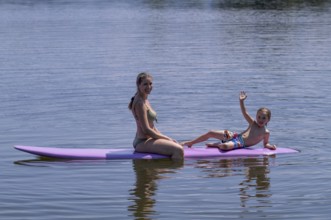 Mother, woman, blonde, in bikini and son, boy, 6 years, sitting on board, waving, surfboard,