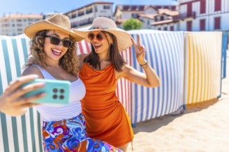 Two cheerful women wearing summer hats and sunglasses are taking a selfie with a smartphone on a
