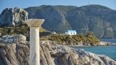 Stone column in front of island with chapel and blue roof, surrounded by azure blue sea, Agios
