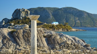 Ancient column and stones in front of island with chapel, blue sky in the background, Agios