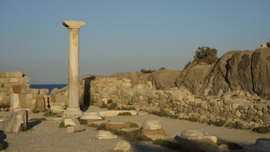 Ancient ruins with a standing stone column and surrounding rock structures in warm sunlight, Agios