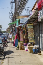 Tourists walk down narrow sidewalk filled with items for sale along Ban Du street in Chiang Rai,