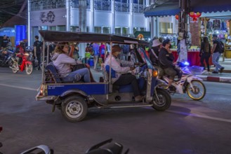 Motor scooter taxi with tourists merges into traffic on a busy downtown street outside the Night