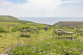 Outdoor cafe with a alone woman sitting by a table on a meadow with a sea view in the summer,