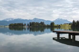 A tranquil lake with a wooden jetty, surrounded by mountains and trees under a cloudy sky