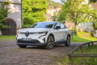 White car parked on a cobbled path in front of old buildings and a green summer backdrop, Renault