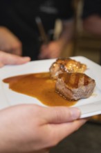 Hand holding a plate with two pieces of roasted meat and plenty of sauce in a kitchen, event and