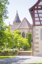 Church tower towers over green trees next to a half-timbered building in summer weather, event and