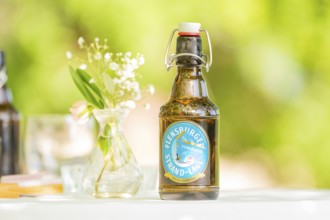 Bottle of Flensburger Strand beer on a table next to a vase of flowers in a green, natural setting,