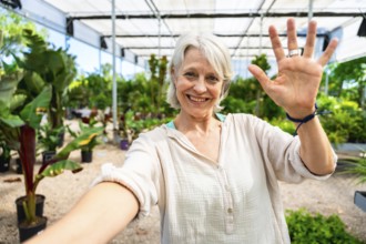 Happy gardener waving at the camera while taking a selfie, surrounded by plants in a greenhouse,