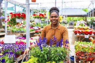 Smiling african american woman pushing shopping cart full of flowers and plants in a greenhouse