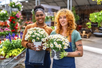 Two female garden center workers holding pots of white daisies smiling in a greenhouse or plant