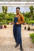 Young african american woman, dressed in denim overalls, posing confidently in a greenhouse,
