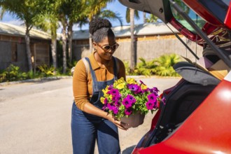Young woman gardener carefully placing colorful petunia flower pots into the trunk of her red car