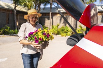 Happy senior woman carrying colorful petunias in a pot to the open trunk of her red car, ready for