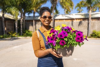 Smiling gardener holding a large pot filled with vibrant, colorful flowers, surrounded by a sunny