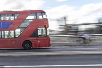 Red double decker bus driving on a london road with a cyclist passing by in the background,