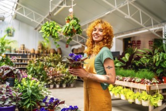 Young saleswoman holding a vibrant potted plant and smiling brightly in a lush greenhouse filled