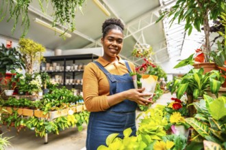 Young woman working in a plant shop, holding a potted flower and smiling surrounded by many plants