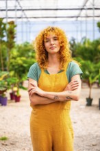 Young woman with curly red hair and tattoos wearing yellow overalls, standing with crossed arms in