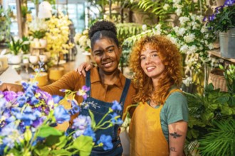 Two happy multiethnic female florists are smiling and posing surrounded by colorful flowers and
