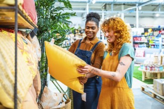 Two cheerful sales assistants collaborating in a home decor store, holding a vibrant yellow pillow