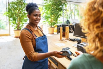 Smiling cashier holding pos terminal while customer paying with smartphone using nfc technology in