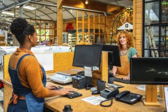 Two young shop assistants are working at the checkout of a plant nursery, using computers and a
