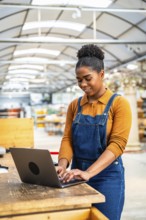 Young woman wearing denim overalls typing on laptop in plant nursery, managing inventory and online