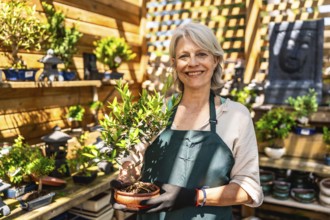 Senior gardener woman smiling while holding a small bonsai tree in a vibrant shop, showcasing her