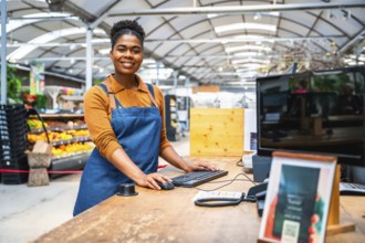 Young female cashier smiling and working at cash register in supermarket with fresh produce in