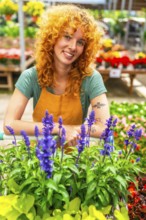 Smiling redhead gardener standing behind a cart overflowing with vibrant purple flowers, surrounded