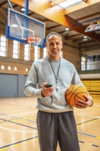 Mature basketball coach holding a ball and stopwatch, smiling confidently in an empty gymnasium,