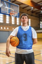 Young male basketball player holding ball in indoor court posing after training, looking confident