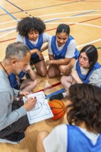 Basketball coach explaining game strategy to female athletes on a clipboard, during a time out in a