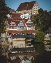 Half-timbered houses with red roofs are reflected in the river next to a wooden bridge in a