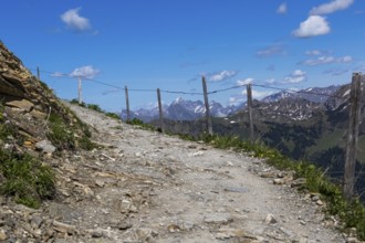 Rocky path with wooden fence, winding through a rugged, alpine mountain landscape under a blue sky