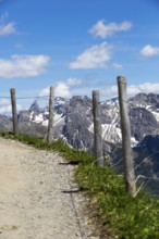 A path lined with wooden fences leads through an alpine landscape with imposing mountain peaks