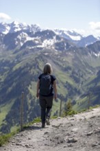 A hiker on a mountain path over green slopes with a view of the vast, snow-covered mountain