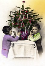 1920, 2 children under a Christmas tree, Two children decorating a small Christmas tree on a table,